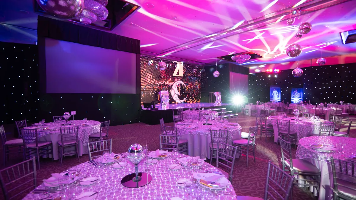 A large room with round tables set for a formal event, featuring white tablecloths, silver chairs, and a stage with a large screen and a pro